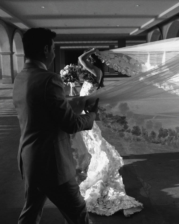 Groom lifting the bride's flowing floral-appliqued veil in a colonnade, black and white