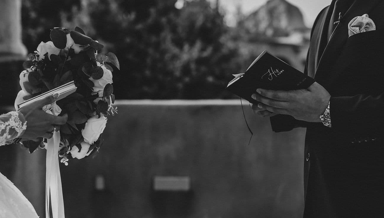 Groom holding a 'His Vows' book across from the bride's bouquet, black and white
