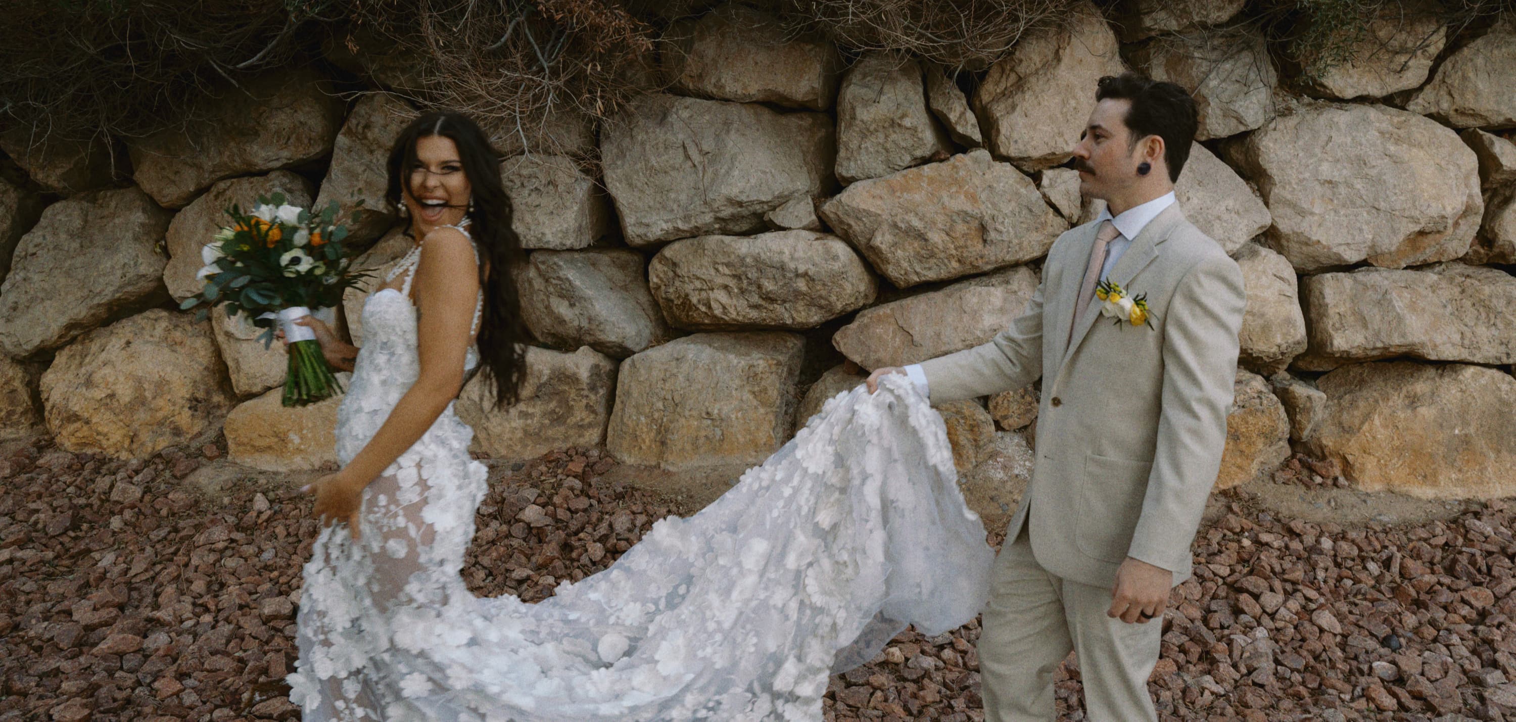 Groom carrying the train of the bride’s lace gown along a stone wall