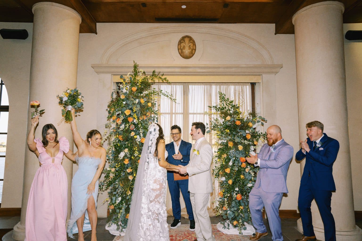Wedding party cheering as the couple holds hands beneath floral arch arrangements