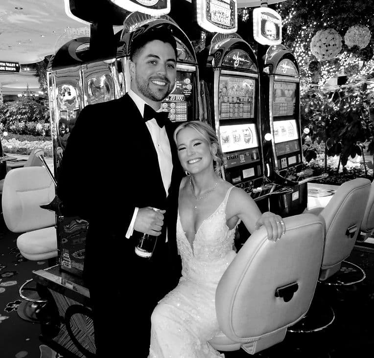 Newlyweds posing with champagne beside casino slot machines, black and white