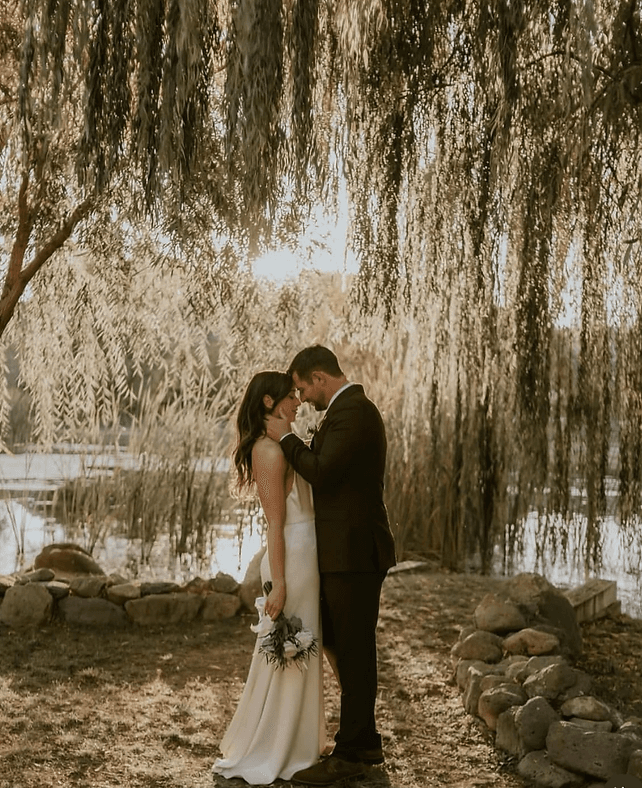 Bride and groom embracing under a willow tree at the water's edge