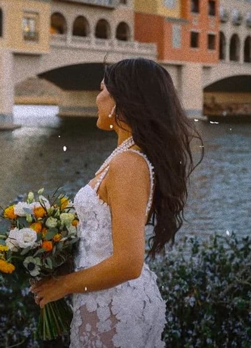 Bride in a lace gown holding an orange and white bouquet beside a canal bridge