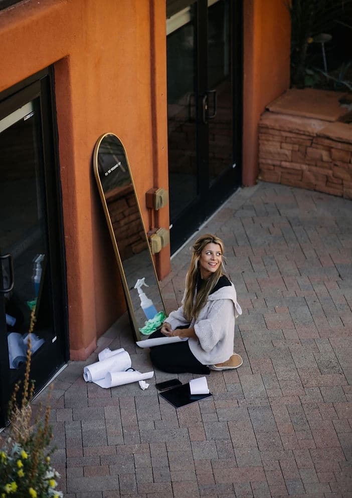 Arabella seated on brick pavement beside a leaning oval mirror with painting supplies