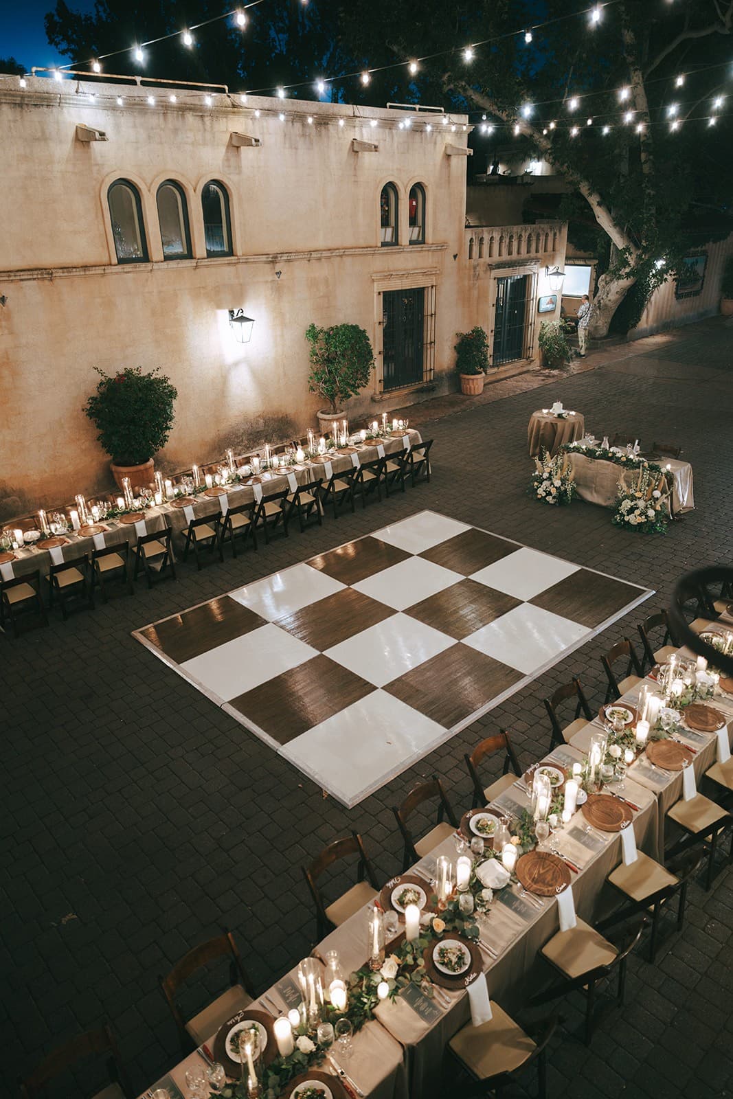 The Calle from above: long candlelit tables with greenery and wood chargers on either side of a checkered dance floor beneath string lights