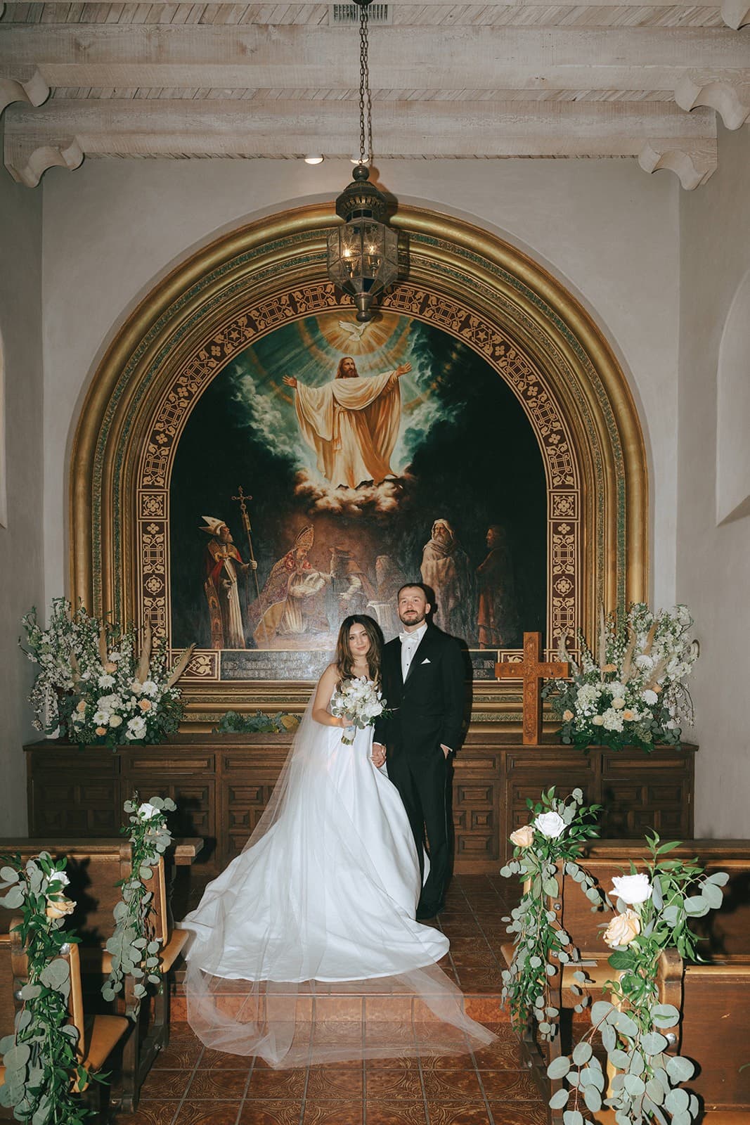 Abby and Connor at the chapel altar beneath the painted arch, white florals on either side
