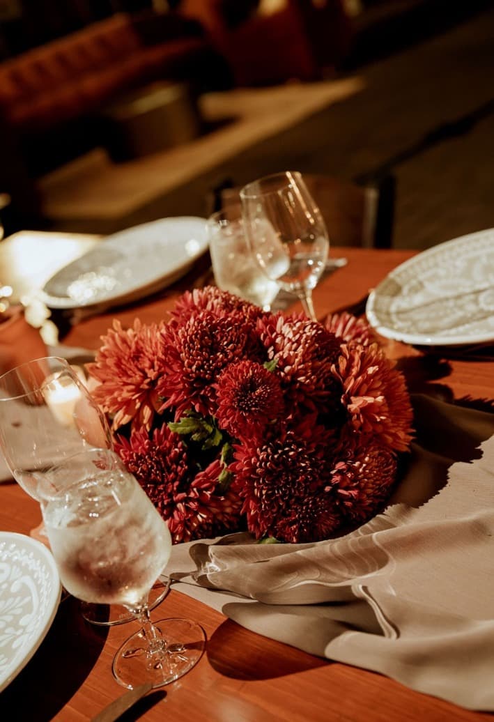 A low arrangement of burnt-red chrysanthemums on a taupe runner between glassware