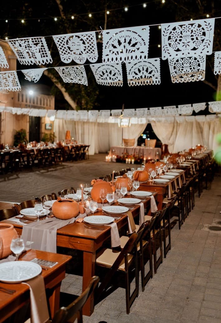 Long wooden banquet tables with terracotta vessels beneath rows of white papel picado and string lights