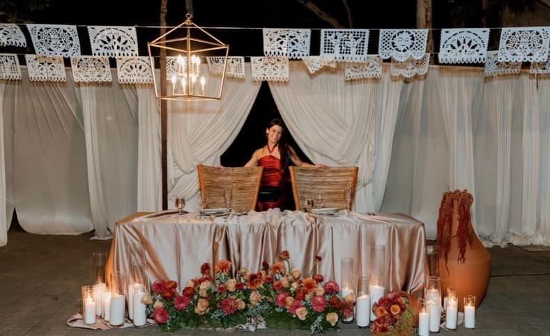 The grooms' sweetheart table with wooden chairs, satin draping, pillar candles and florals beneath papel picado and a lantern