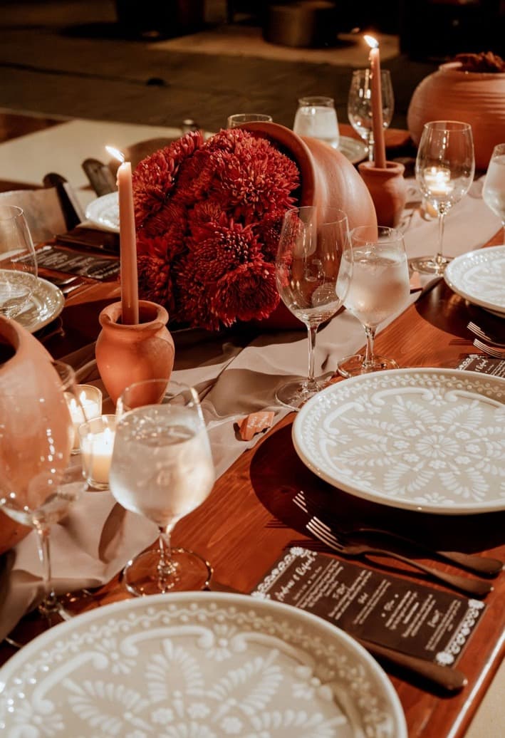 Patterned dinnerware, terracotta candleholders, taper candles and a vessel of red chrysanthemums on a wooden table