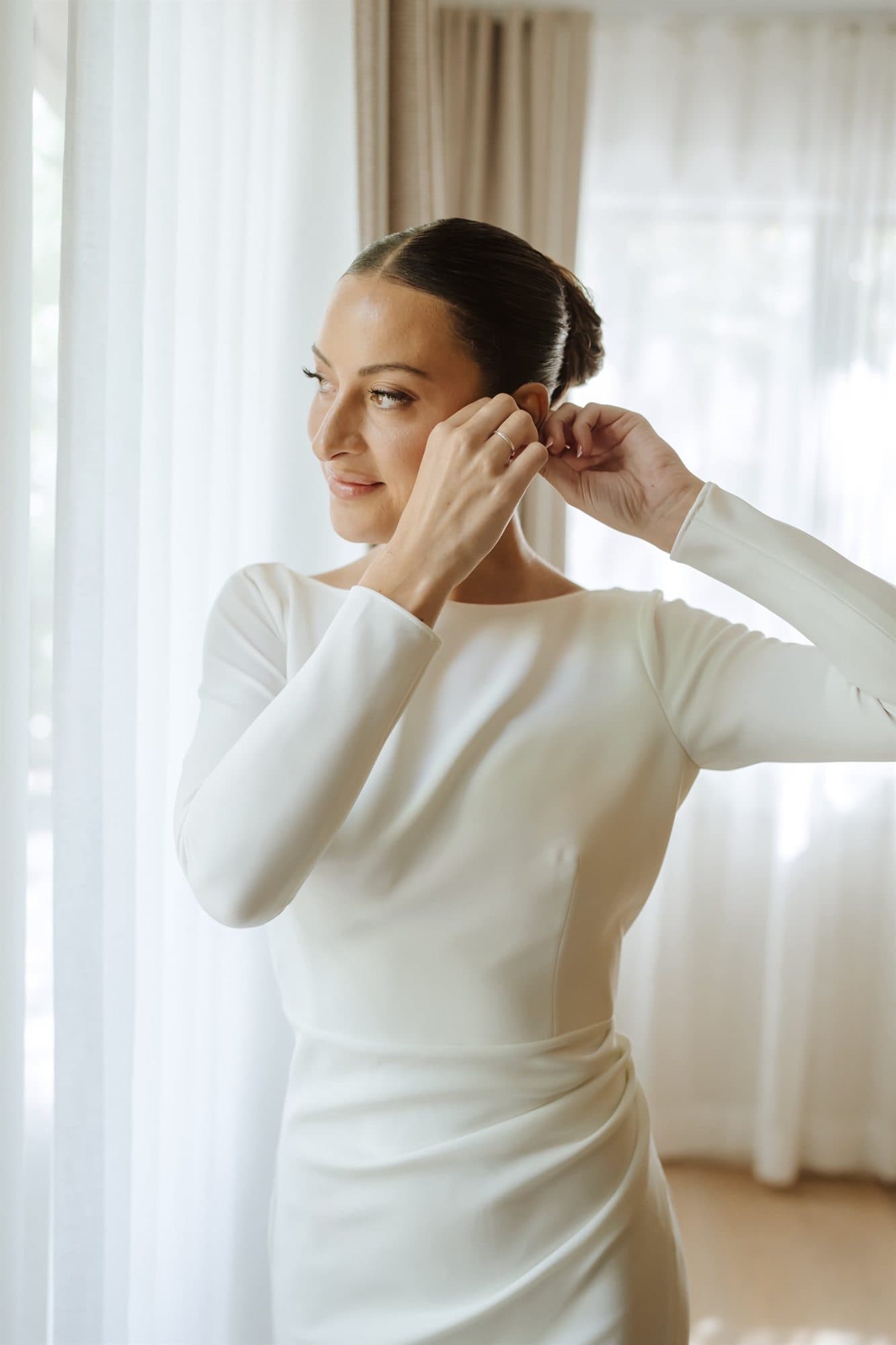 Getting ready by the window in a long-sleeved crepe gown, fastening an earring