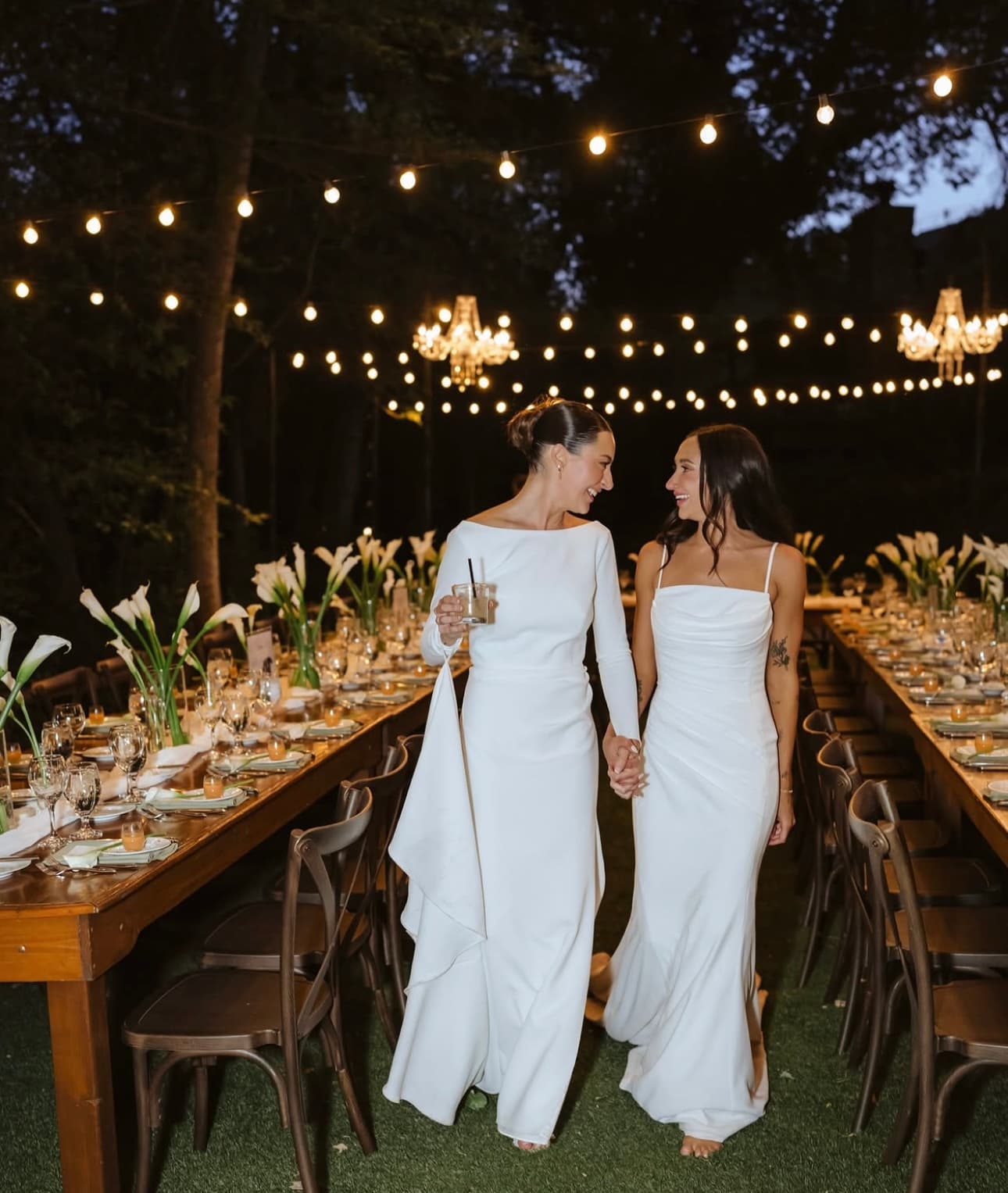 Brynn and Megan walking hand in hand between long calla-lily tables beneath string lights and crystal chandeliers