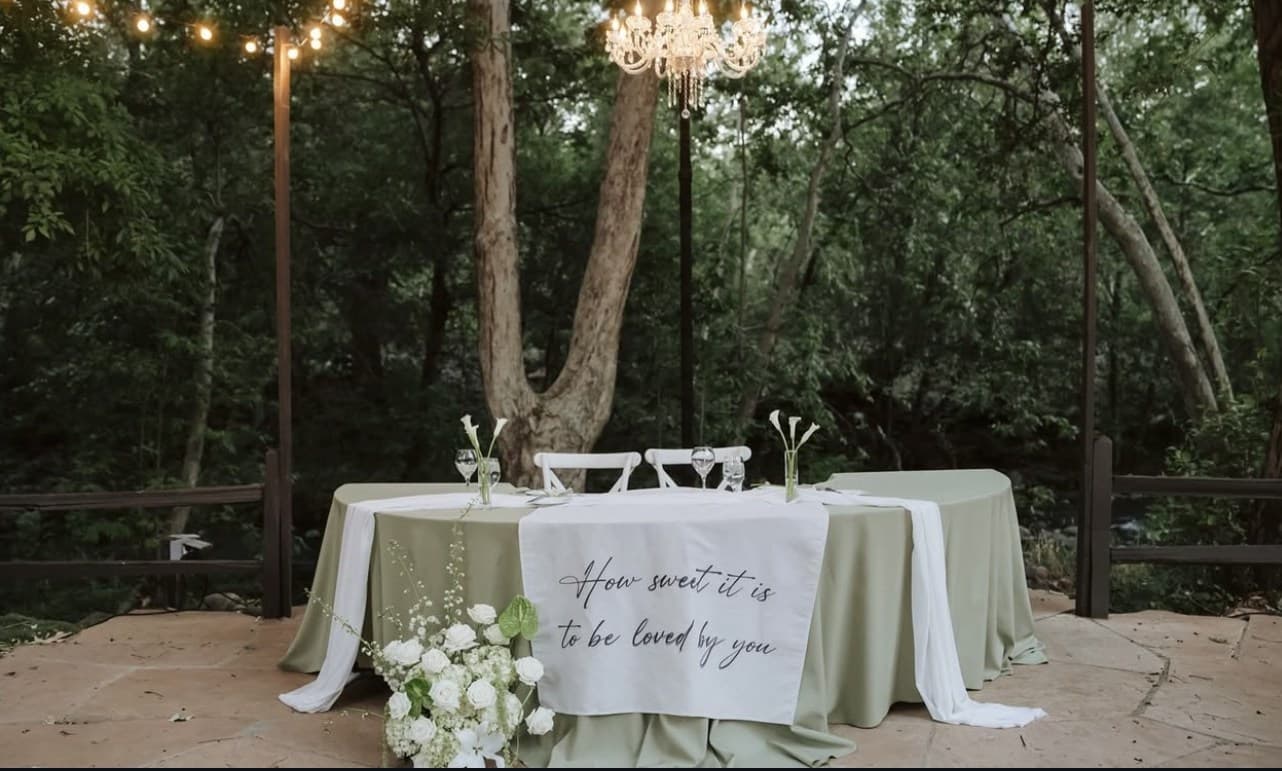 The sweetheart table along Oak Creek beneath a crystal chandelier, with a "How sweet it is to be loved by you" runner