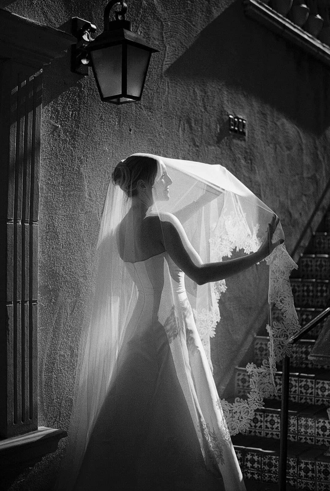 A bride lifting her veil on a shadowed staircase, in black and white
