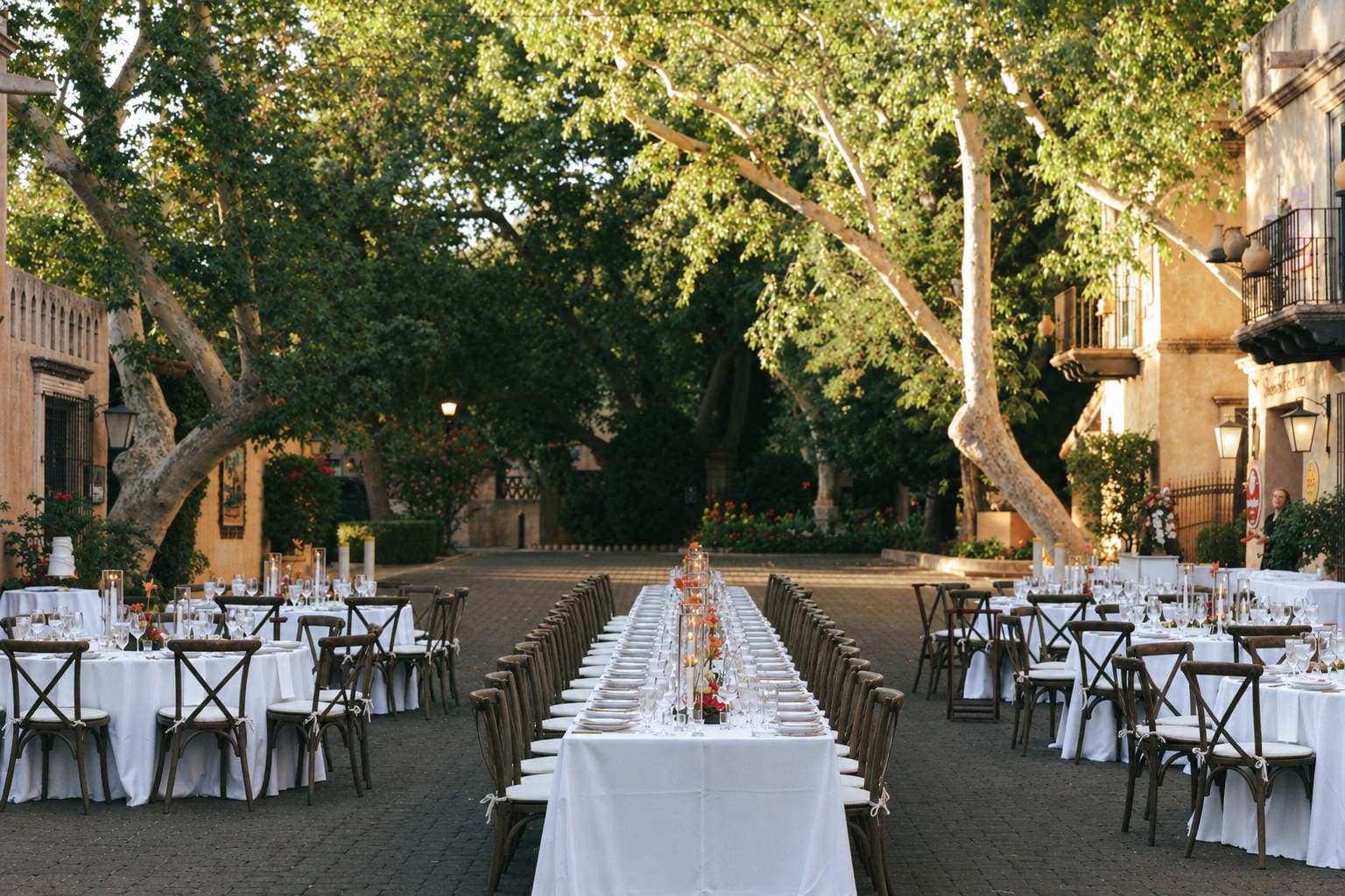 Long white banquet table with tall glass candles and orange florals beneath the sycamores at Tlaquepaque