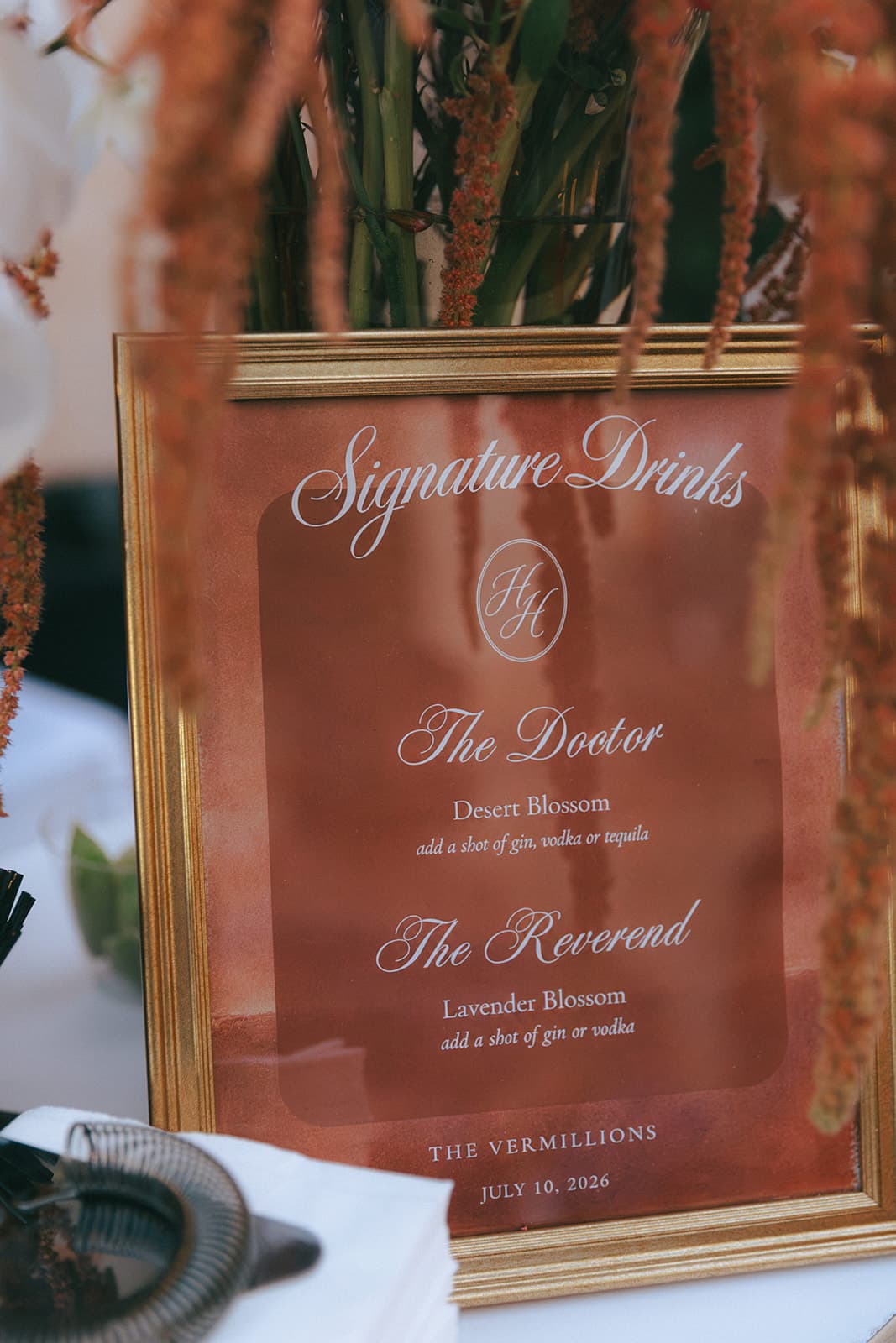 Gold-framed signature drinks sign with the H&H monogram beneath hanging amaranthus