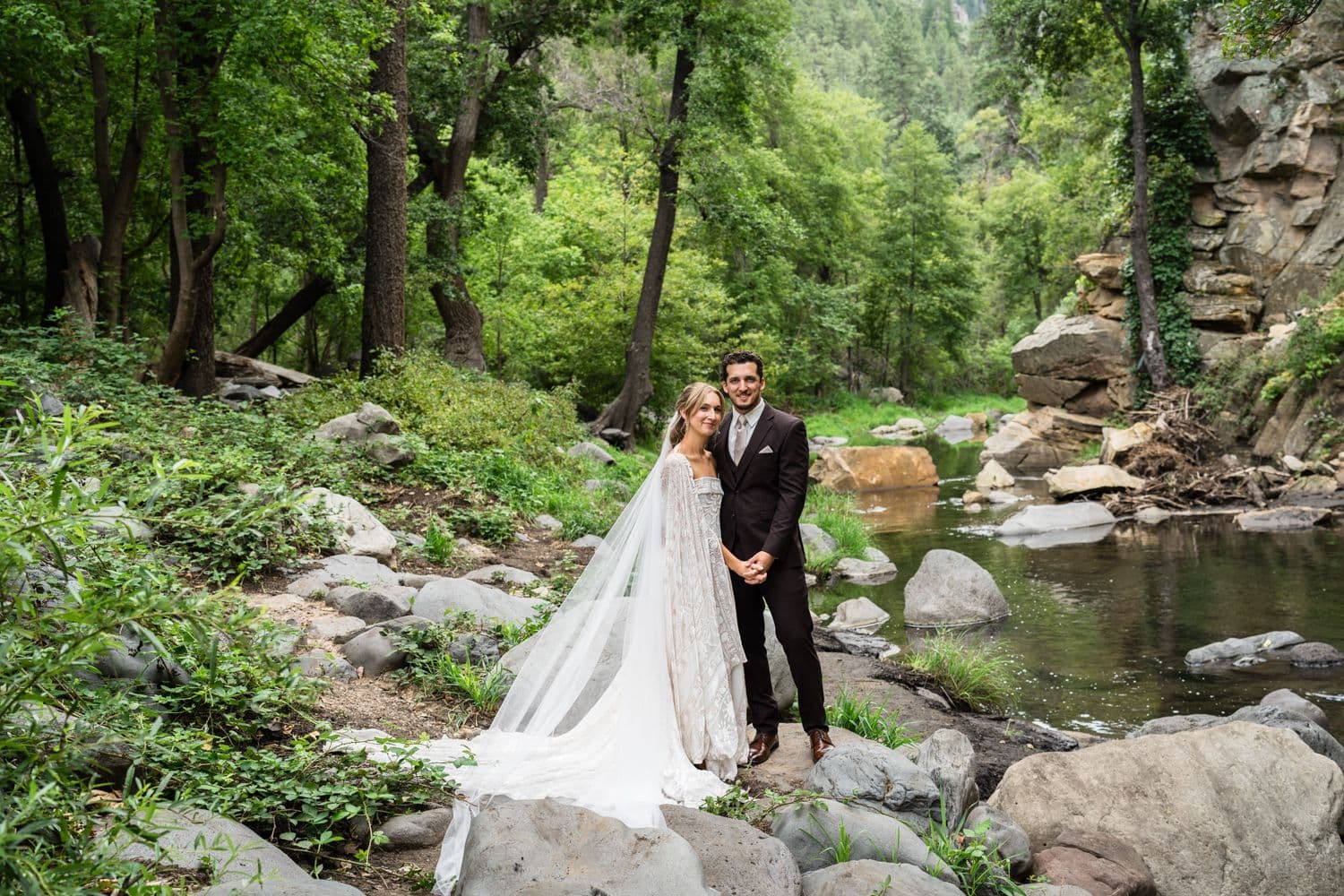 Bride in a long veil and caped lace gown with the groom beside Oak Creek in the forest