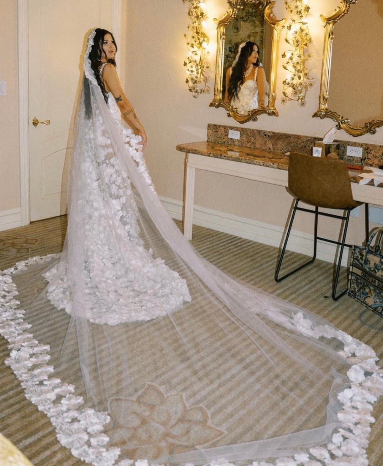 Isa looking back over her shoulder, her floral-edged cathedral veil spread across the floor beside a gilded mirror