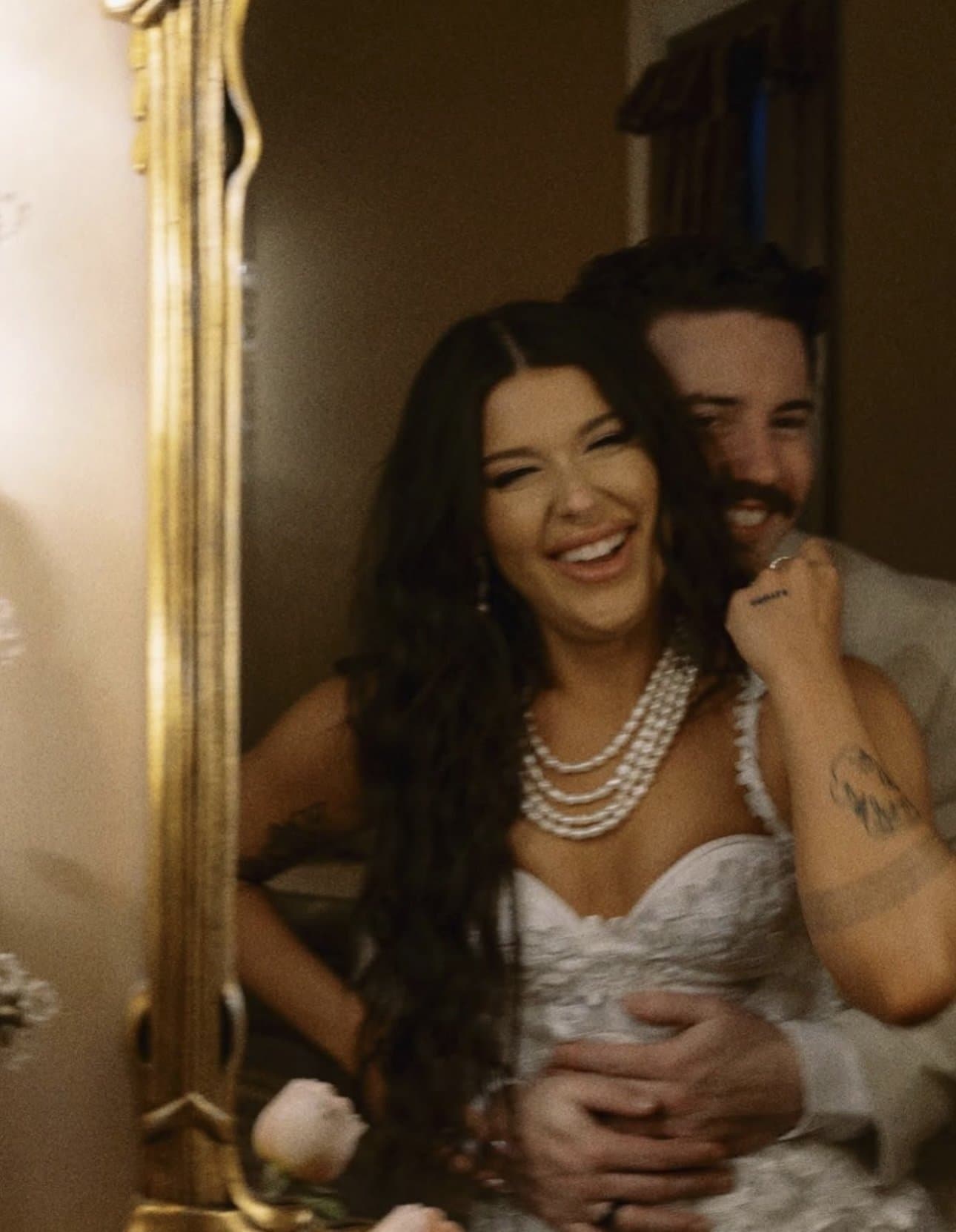 Isa laughing in a gilded mirror as Dylan wraps his arms around her