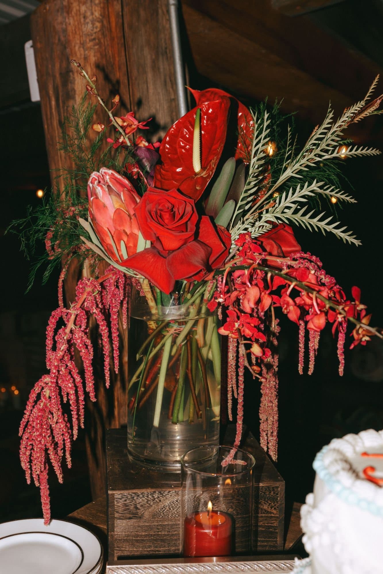 A tall arrangement of anthurium, protea, red roses and hanging amaranthus on a wooden riser