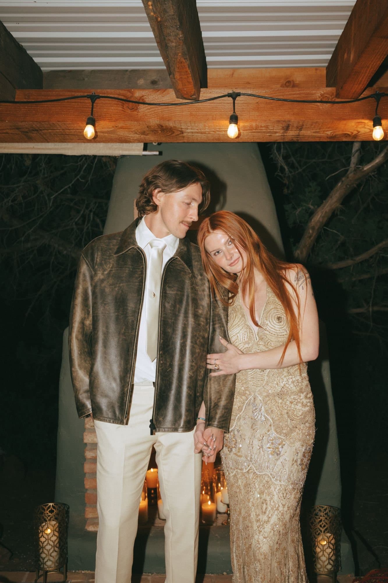 Austin in a leather jacket and Jordan in a beaded gold gown beside a candlelit kiva fireplace