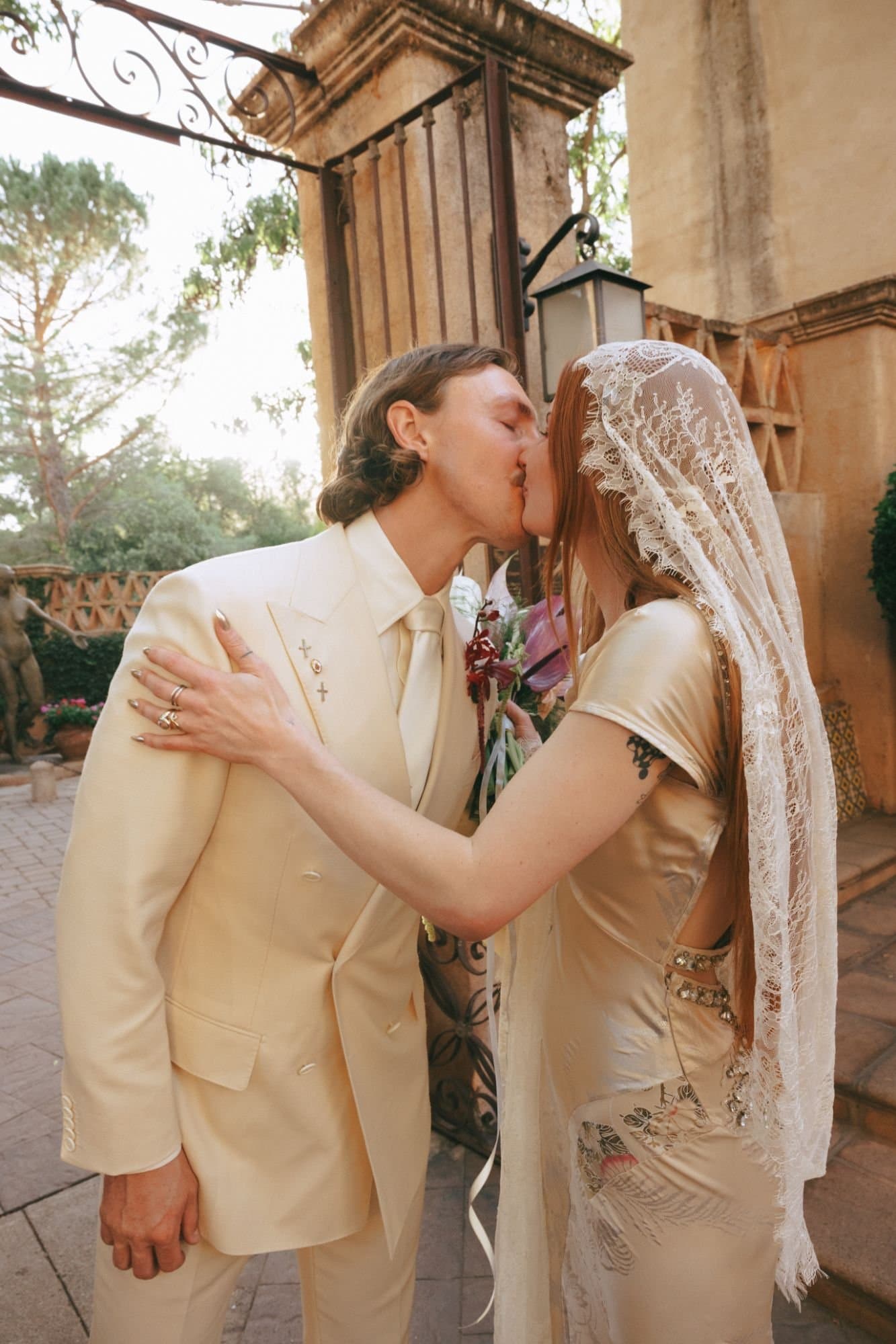 Jordan in a lace mantilla and gold satin gown kissing Austin in an ivory suit at the Tlaquepaque gate