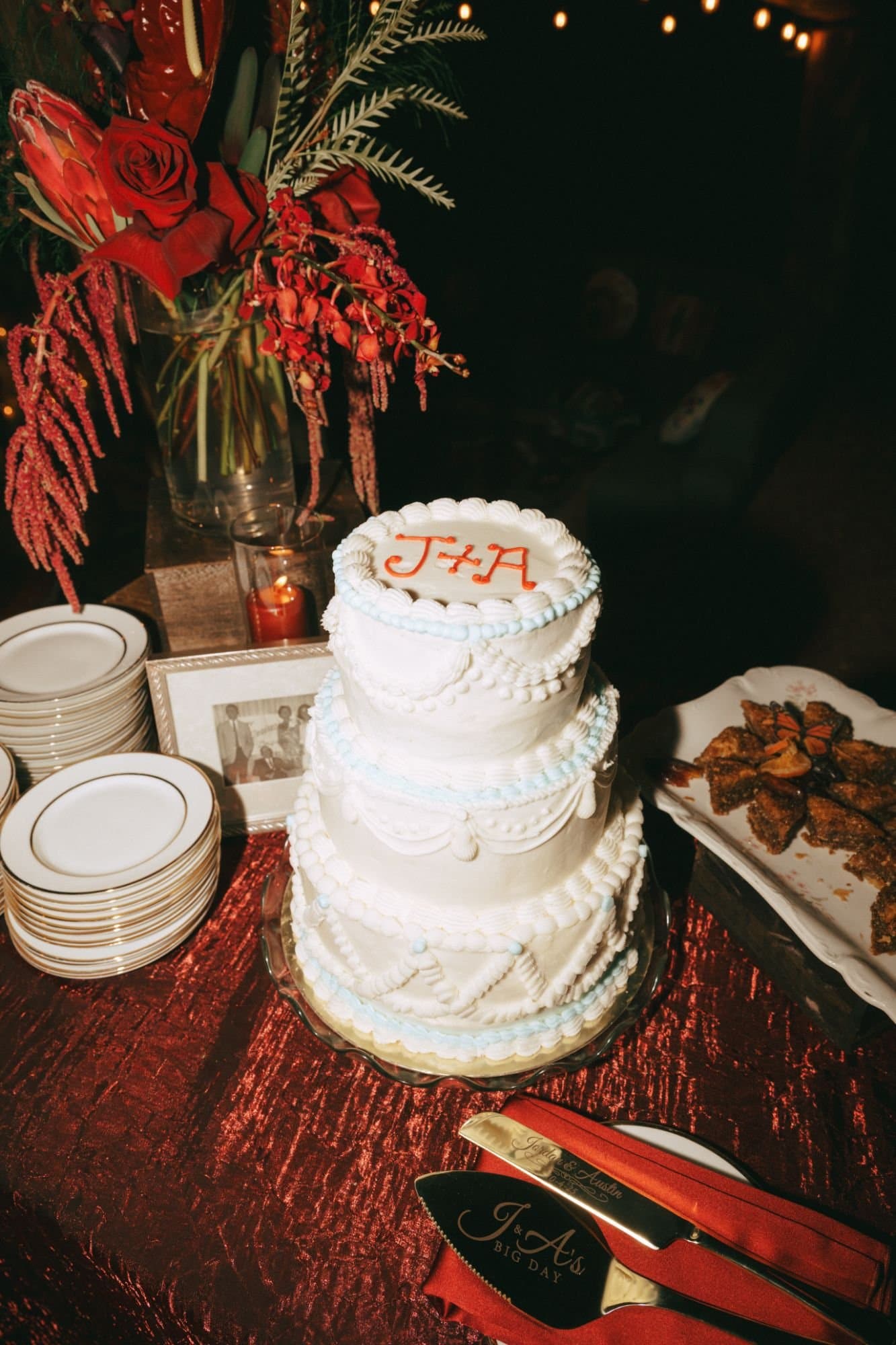 A three-tier vintage piped cake with "J + A" in red beside engraved cake servers and a framed family photo