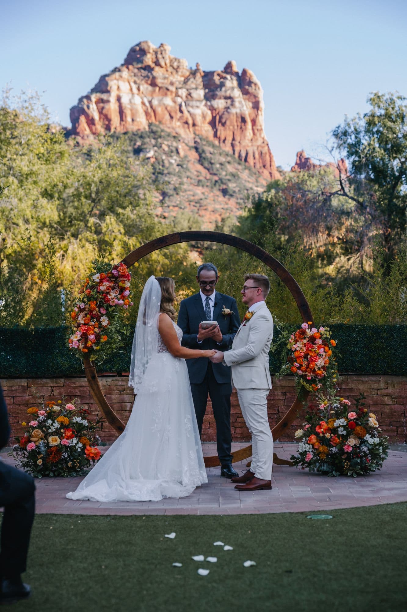 Jenna and Derek holding hands before a circular floral arch with the Sedona red rocks behind them