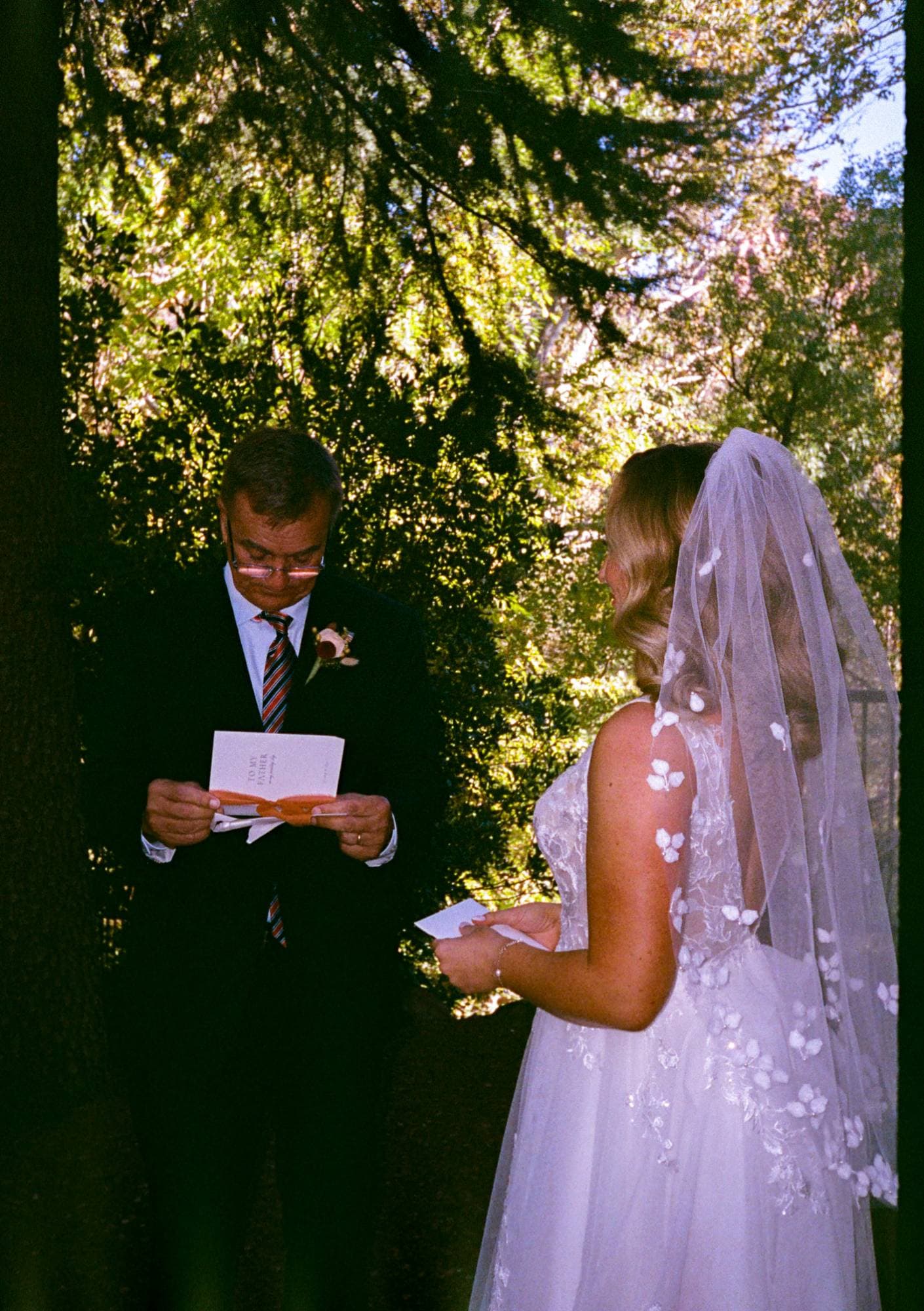 Jenna in her floral veil as her father reads a letter beneath the trees, on film