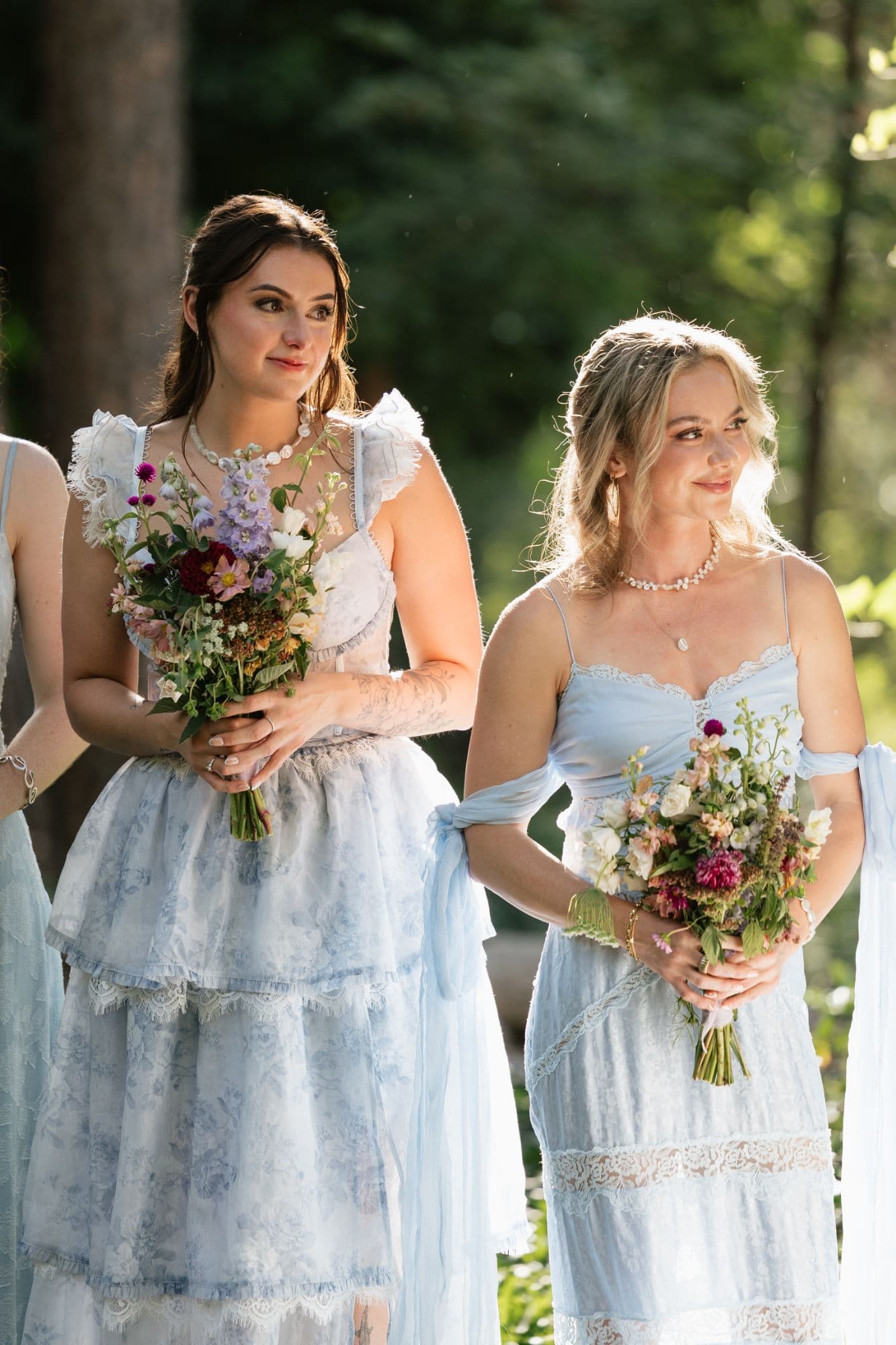 Bridesmaids in soft blue lace holding wildflower bouquets in the sunlit forest