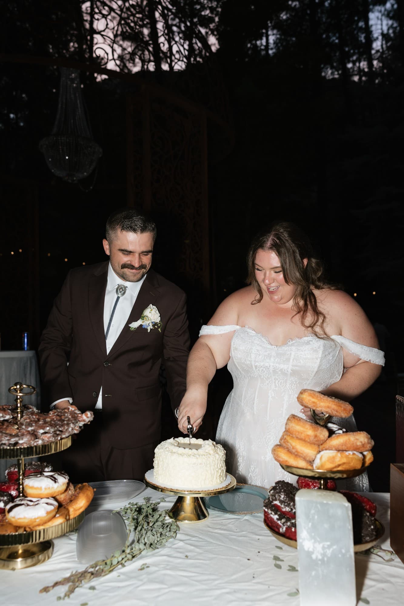 Cutting the cake beneath a crystal chandelier in the forest, surrounded by stacked donuts and selenite