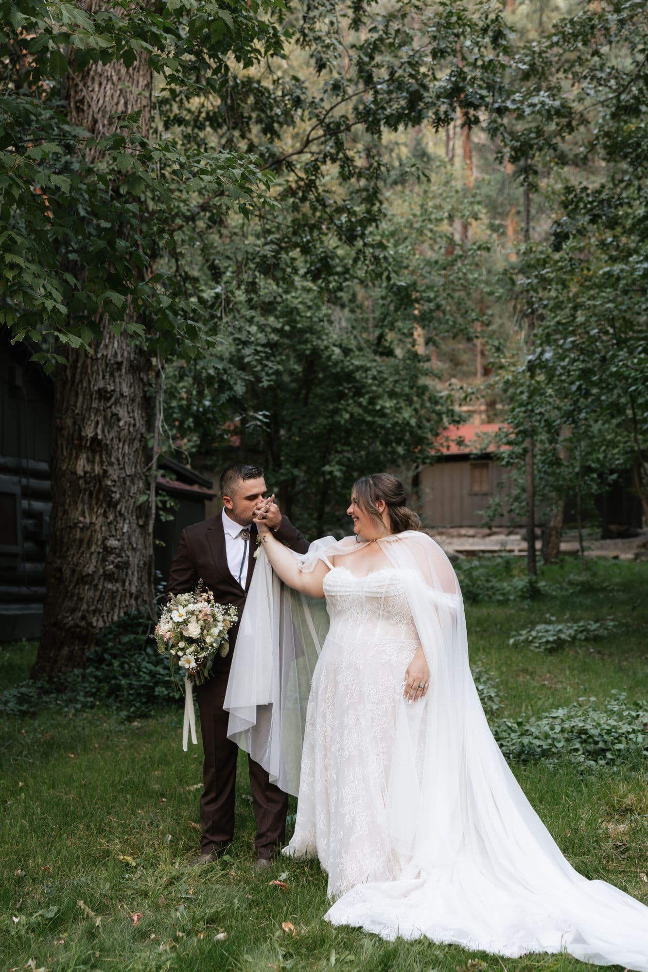 The groom kissing the bride's hand among the trees, her tulle cape trailing across the grass
