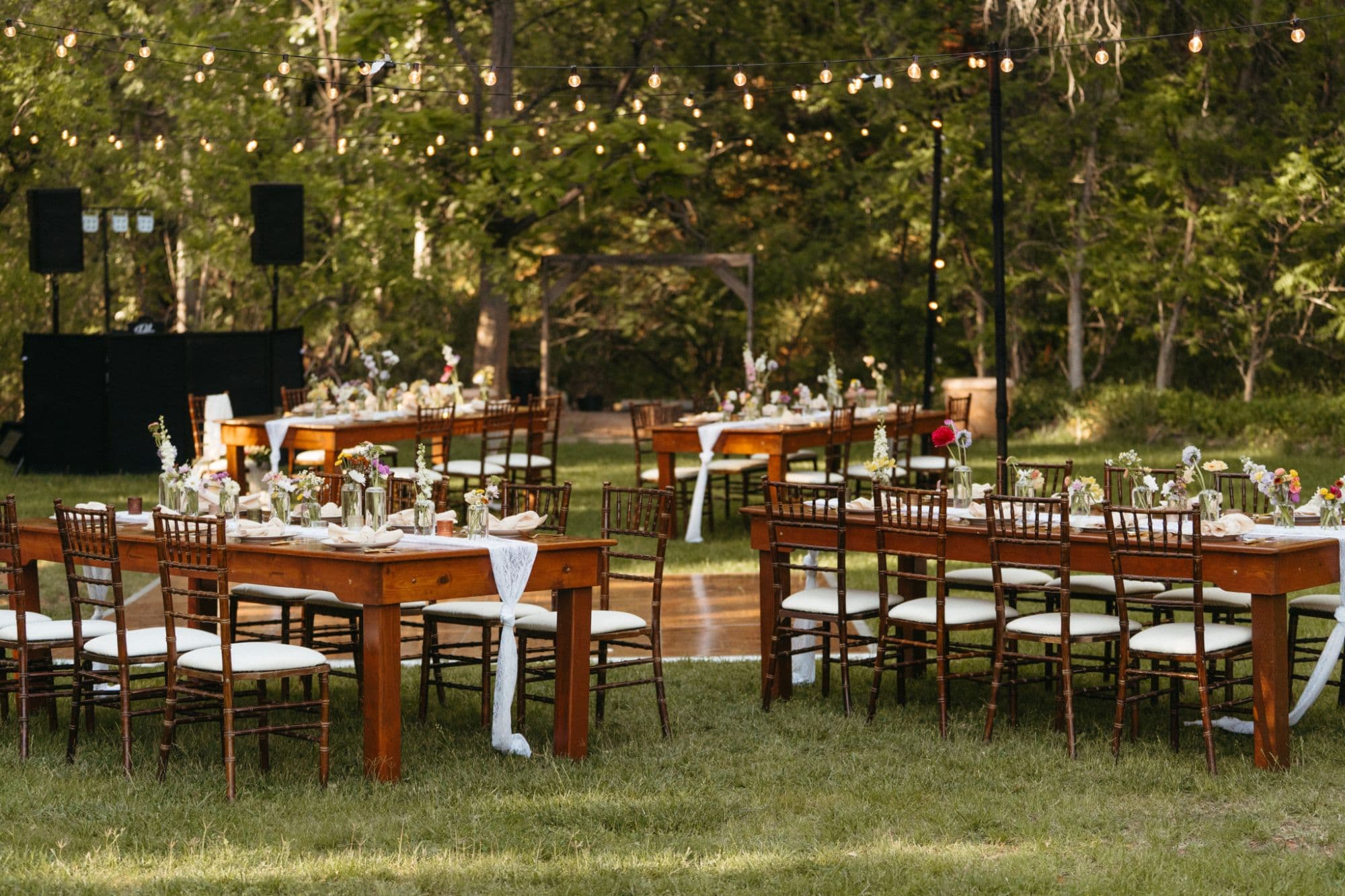 Long wooden garden tables with lace runners and wildflowers in petite glass vases beneath string lights