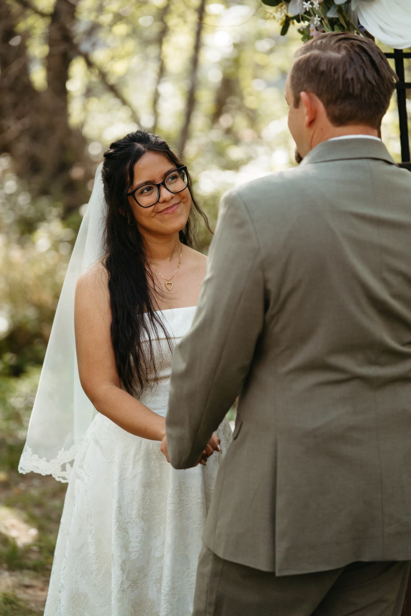 Patricia smiling at Drew as they hold hands for their vows in the sunlit trees