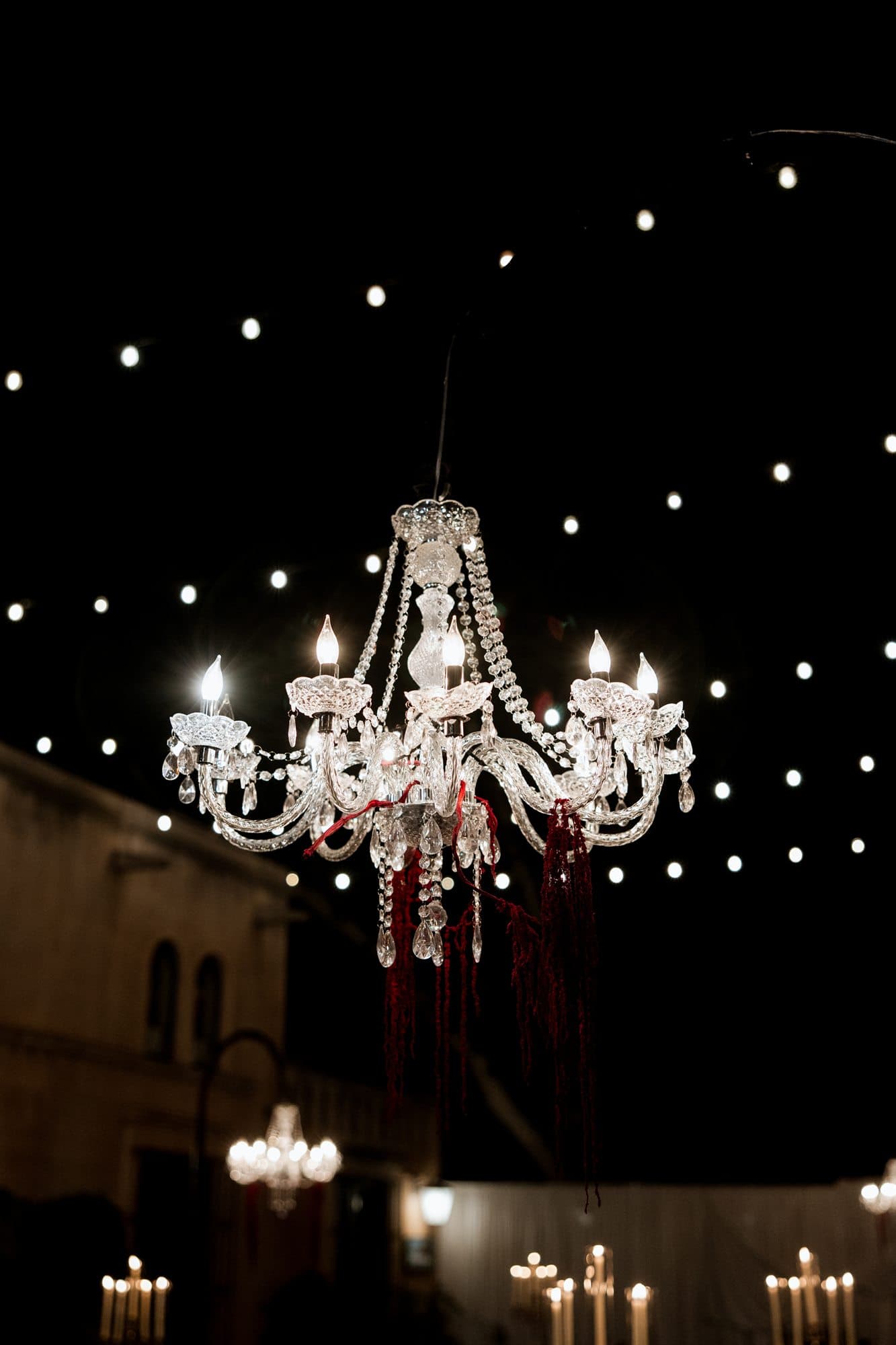 A crystal chandelier dripping with red amaranthus beneath string lights