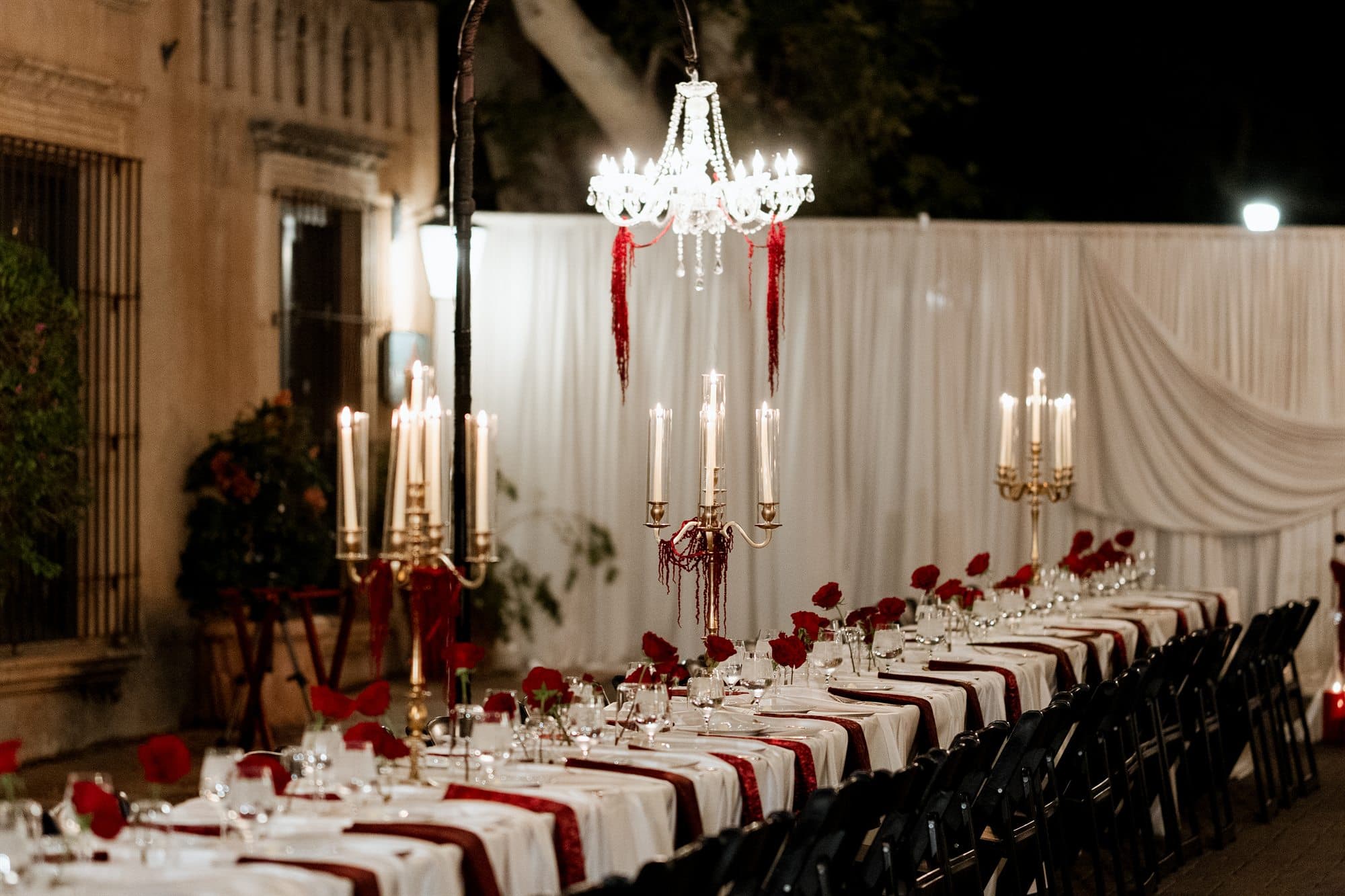 Long banquet tables with burgundy runners and gold candelabras before ivory draping and a crystal chandelier