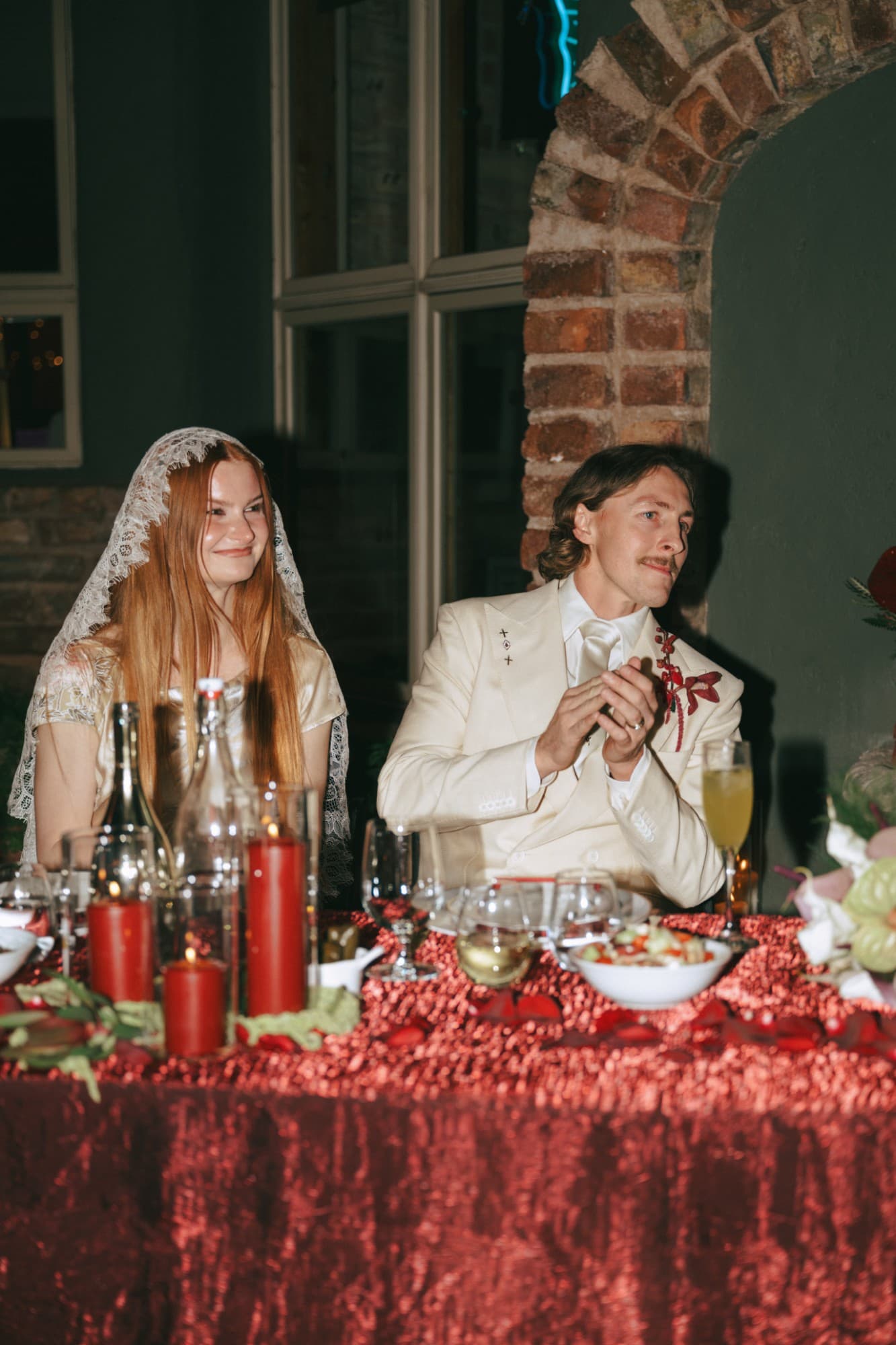 Bride in a lace mantilla and groom in an ivory suit at a red sweetheart table