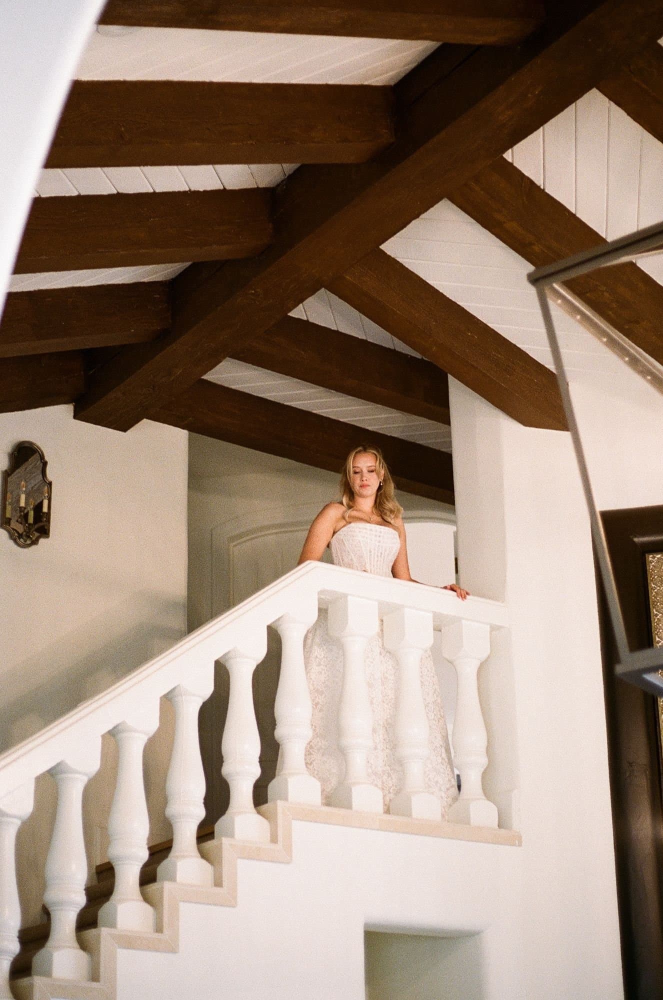 Sophia looking down from a white balustrade staircase beneath dark wood beams