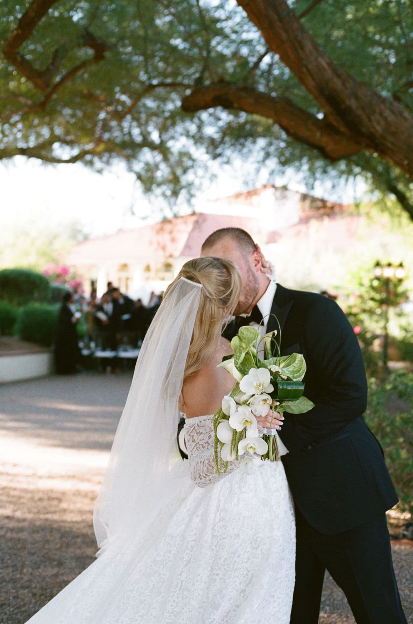 A kiss beneath the mesquite branches as guests look on, her orchid and anthurium bouquet raised