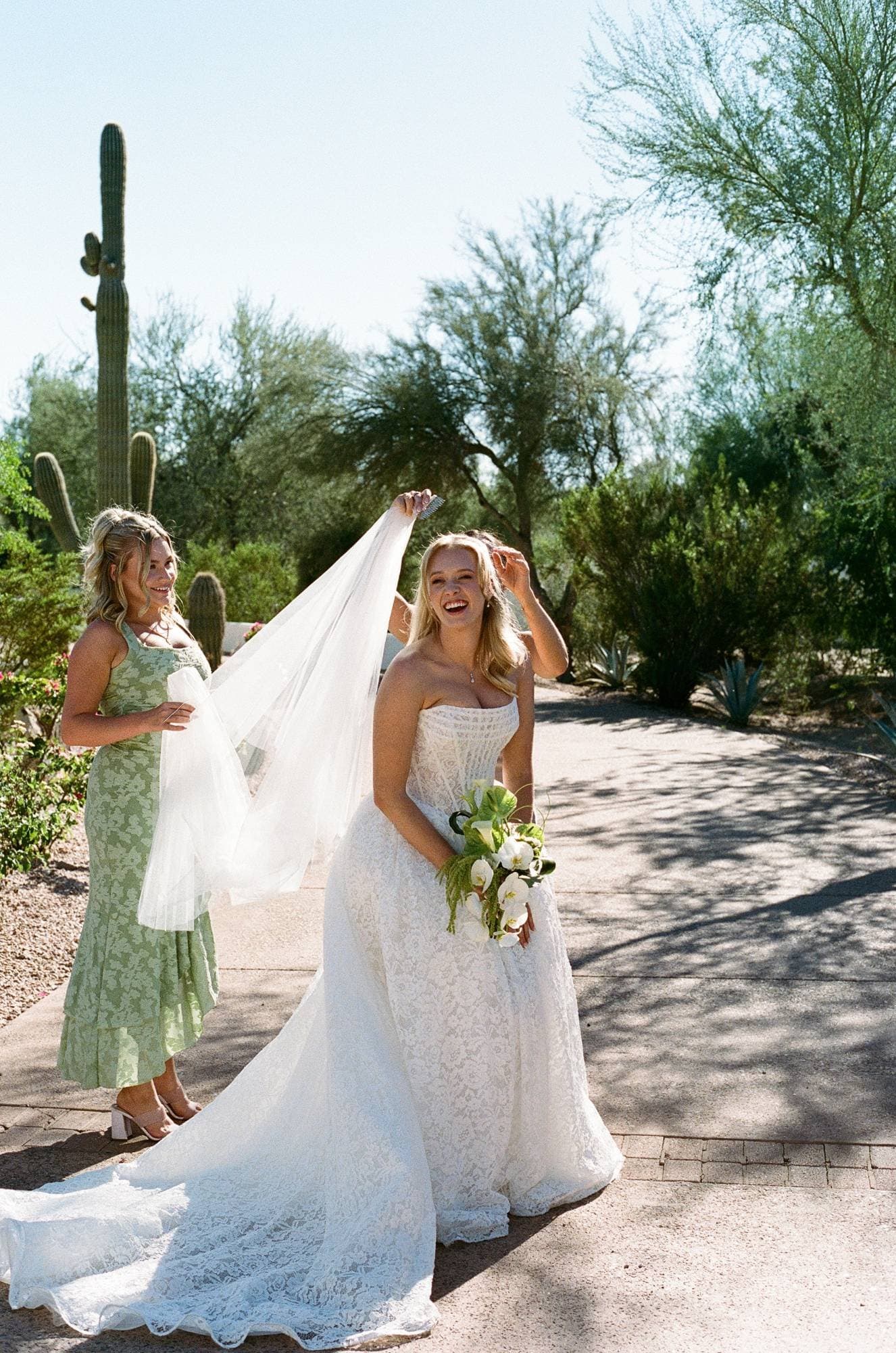A bridesmaid lifting Sophia's veil beside a towering saguaro, her bouquet of orchids, calla lilies and amaranthus in hand