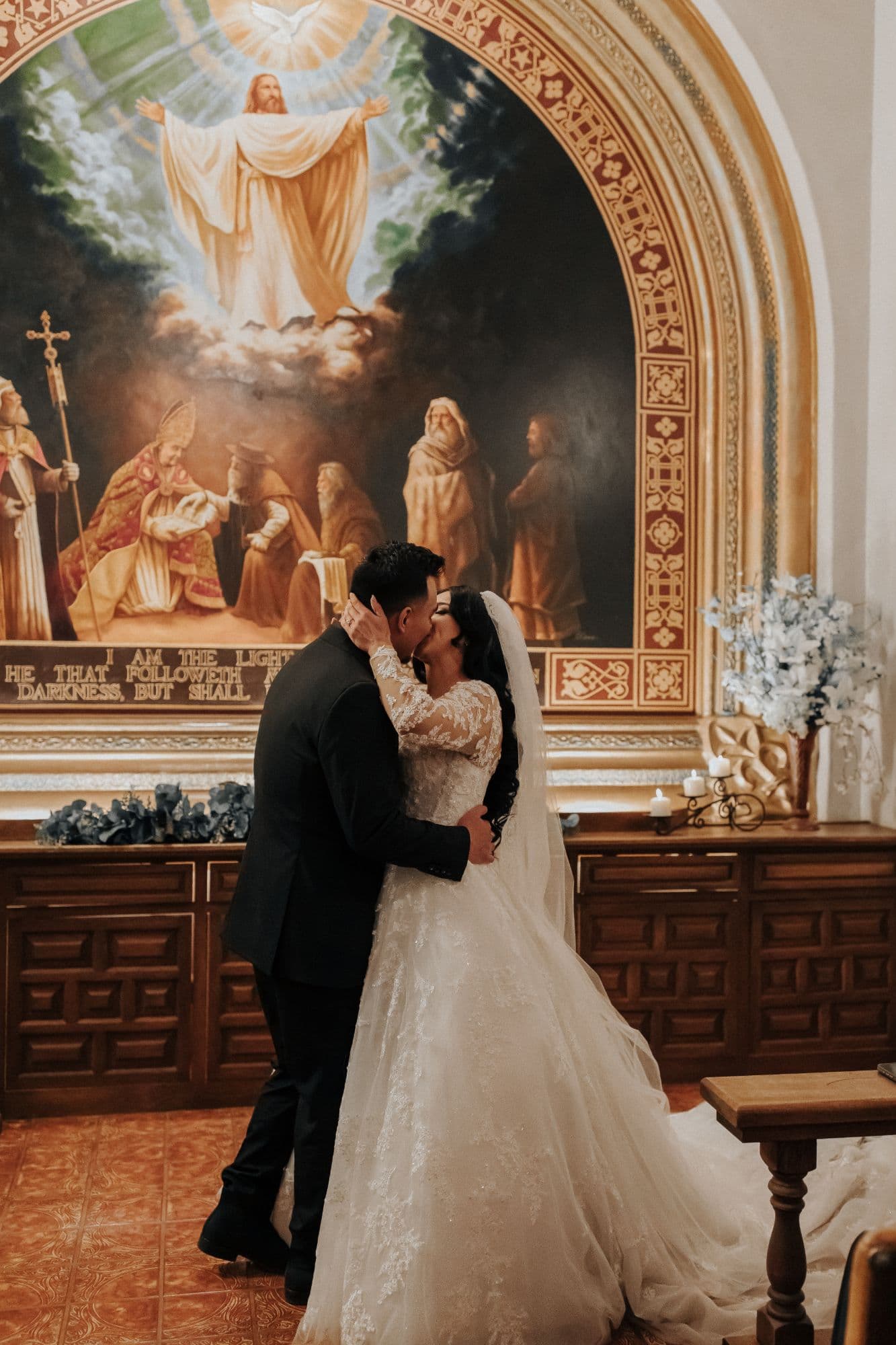 Bride in a long lace-trimmed veil kissing the groom before the painted altar of the Tlaquepaque chapel
