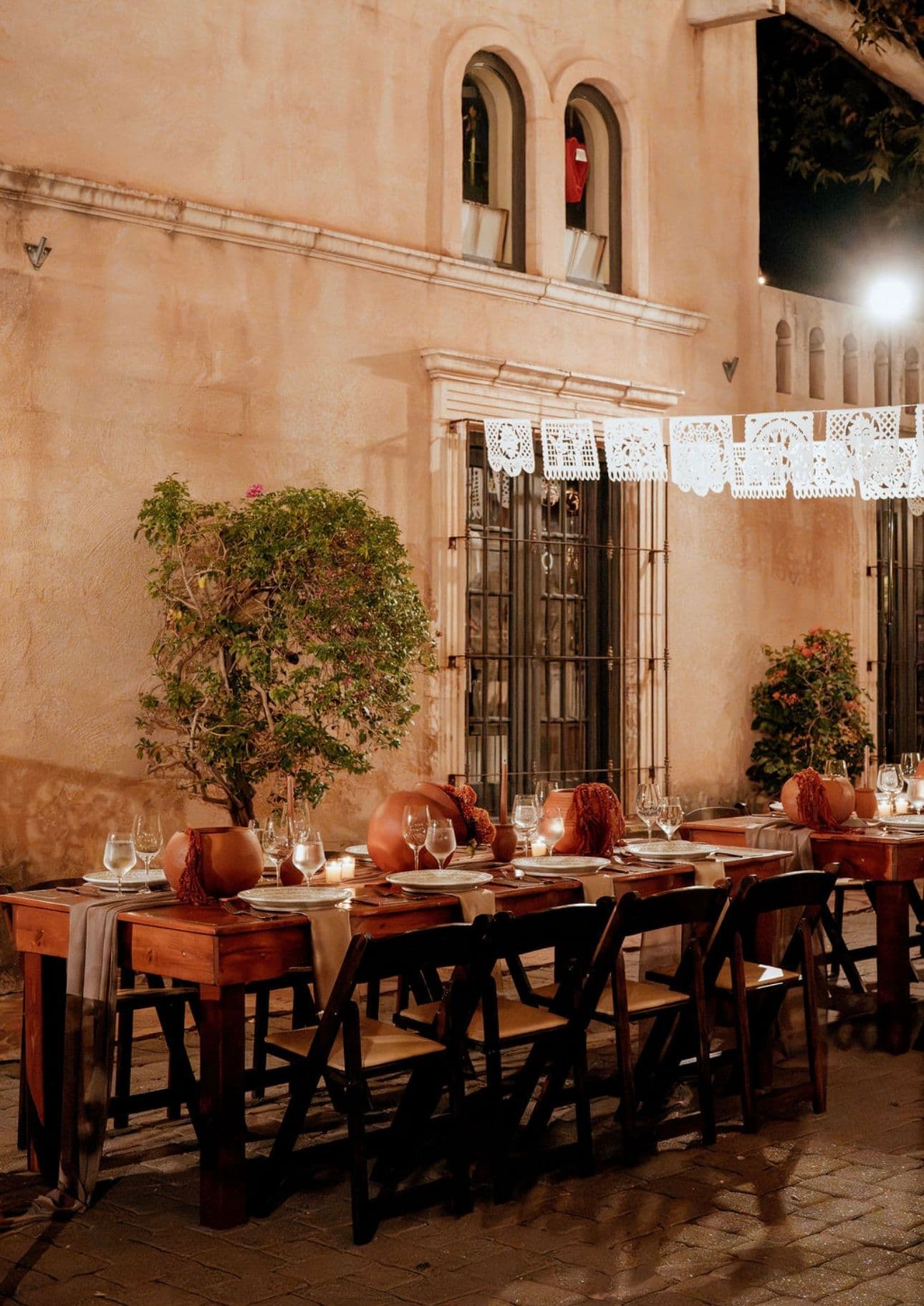 Wooden farm tables set with terracotta vessels and candles beneath papel picado along a Tlaquepaque wall at night