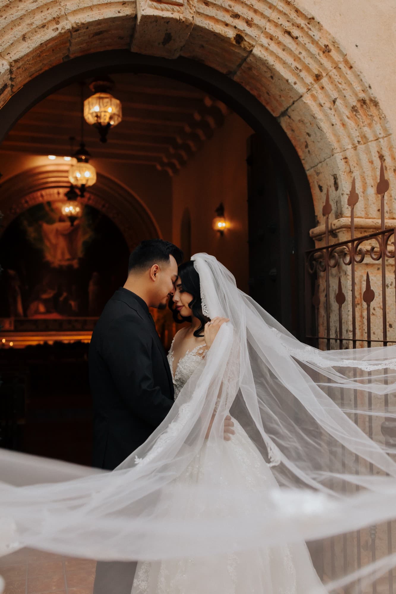 Bride and groom forehead to forehead beneath a stone arch as her cathedral veil sweeps across the frame